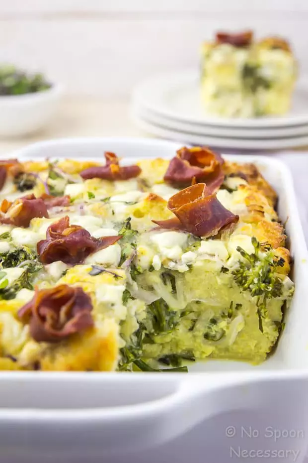 Straight on shot of an Overnight Broccolini & Goat Cheese Strata topped with crispy prosciutto in a white baking dish with a square slice of strata missing from the dish and placed on a plate blurred in the background. 