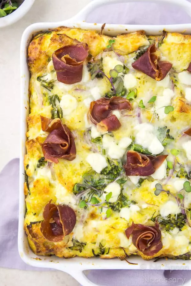 Overhead, up-close shot of an Overnight Broccolini & Goat Cheese Strata topped with crispy prosciutto in a white baking dish on top of a light purple linen with a ramekin of microgreens tucked in the corner of the shot.