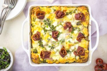 Overhead shot of Overnight Broccolini & Goat Cheese Strata topped with crispy prosciutto in a white baking dish on top of a light purple linen with plates, forks, vine ripe tomatoes, and a ramekin of microgreens surrounding the baking dish.
