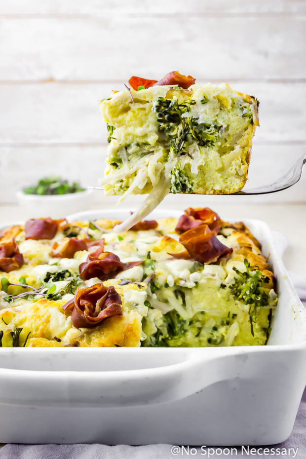 Straight on shot of a slice of Overnight Broccolini & Goat Cheese Strata topped with crispy prosciutto being lifted with a spatula out of a white baking dish with a small bowl of microgreens blurred in the background.