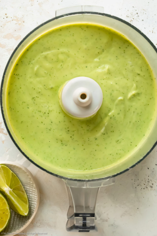 Overhead photo of homemade creamy avocado dressing in the bowl of a food processor with a ramekin of fresh lime wedges next to the bowl.