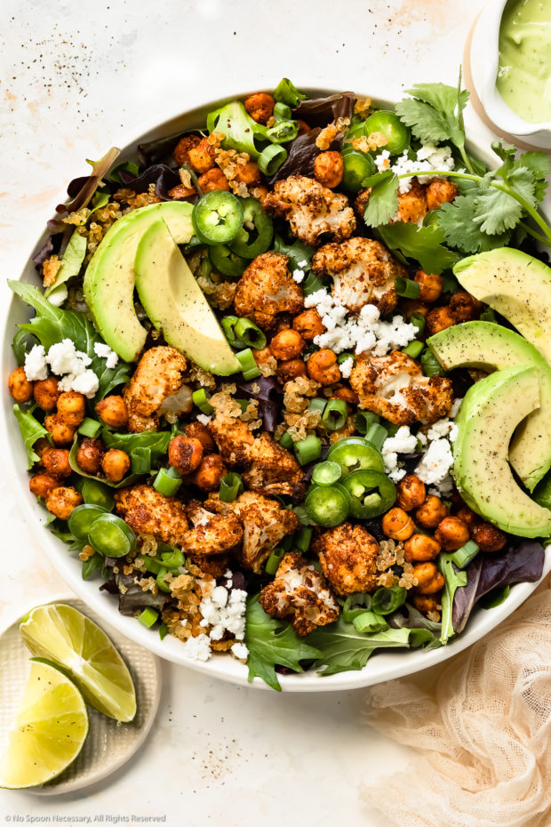 Overhead photo of Roasted Cauliflower Salad with chickpeas, crispy quinoa and avocado in a large white serving bowl with a ramekin of fresh lime wedges and a jar of avocado dressing next to the bowl.