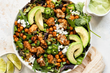 Overhead, landscape photo of Roasted Cauliflower Salad with chickpeas, crispy quinoa and avocado in a large white serving bowl with a ramekin of fresh lime wedges and a jar of avocado dressing next to the bowl.