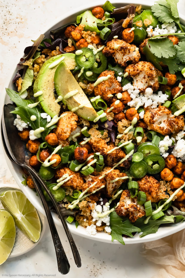 Overhead photo of Roasted and Chickpea Cauliflower Salad drizzled with healthy avocado dressing in a large white serving bowl with two serving spoons inserted into the salad and fresh lime wedges next to the bowl.