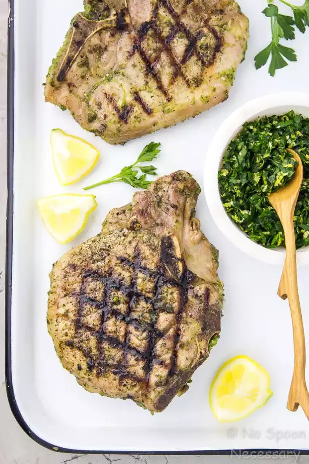 Overhead shot of two grilled Garlic & Herb Pork Chops on a white tray with a ramekin of Gremolata and lemon wedges.