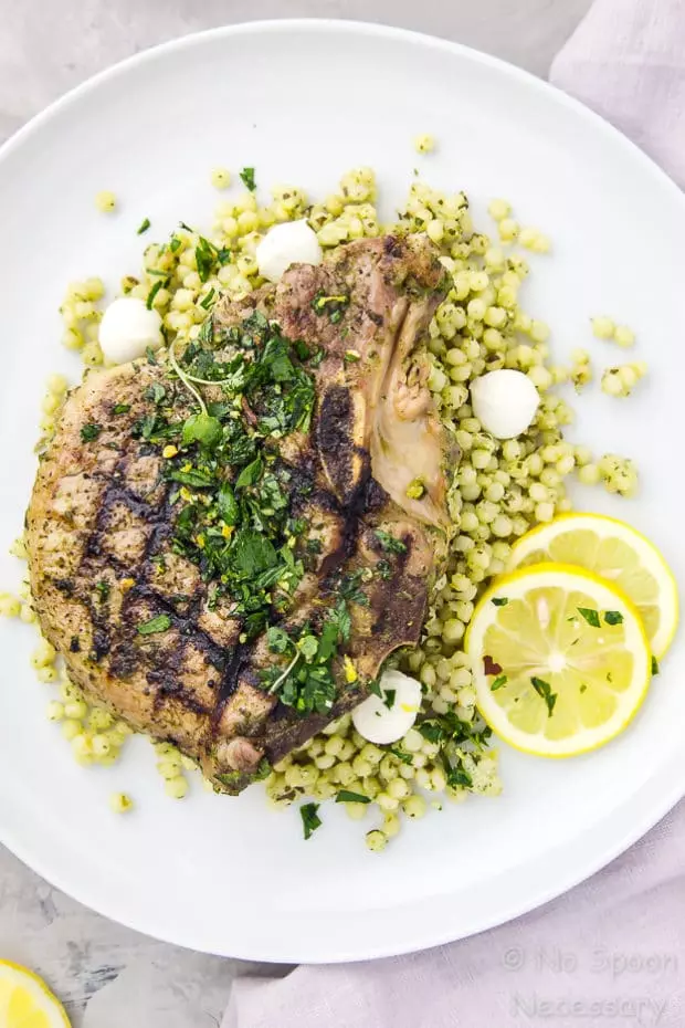 Overhead shot of a white plate containing grilled Garlic & Herb Pork Chop topped with Gremolata on a couscous salad with a purple linen under the plate.