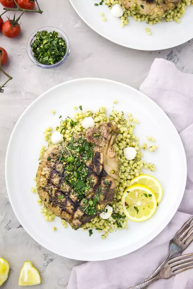 Overhead shot of a white plate containing grilled Garlic & Herb Pork Chop topped with Gremolata on a couscous salad with a purple linen, forks, vine ripe tomatoes, and bowl of gremolata surrounding the plate.