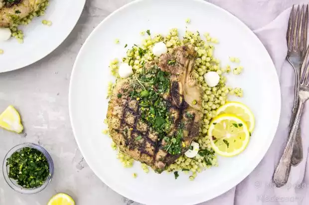 Overhead shot of a white plate containing grilled Garlic & Herb Pork Chop topped with Gremolata on a couscous salad with a purple linen, forks, and bowl of gremolata surrounding the plate.