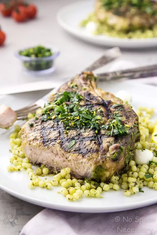 Angled shot of a white plate containing a cut into grilled Garlic & Herb Pork Chop topped with Gremolata on a couscous salad with another plate of chops, vine ripe tomatoes, and bowl of gremolata blurred in the background.