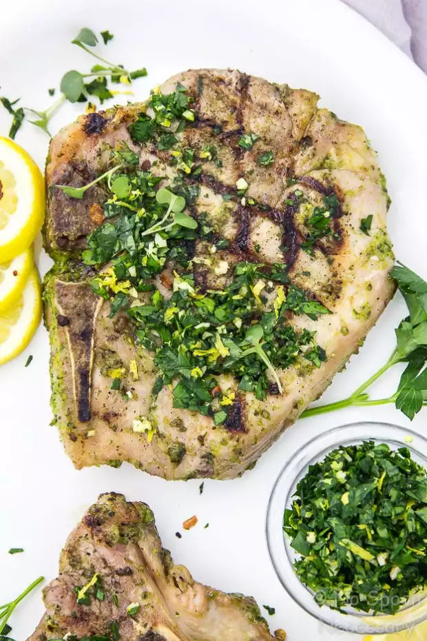 Overhead, up close shot of a grilled Garlic & Herb Pork Chop topped with Gremolata on a white plate with a ramekin of gremolata, fresh parsley and lemon slice surrounding the chop.