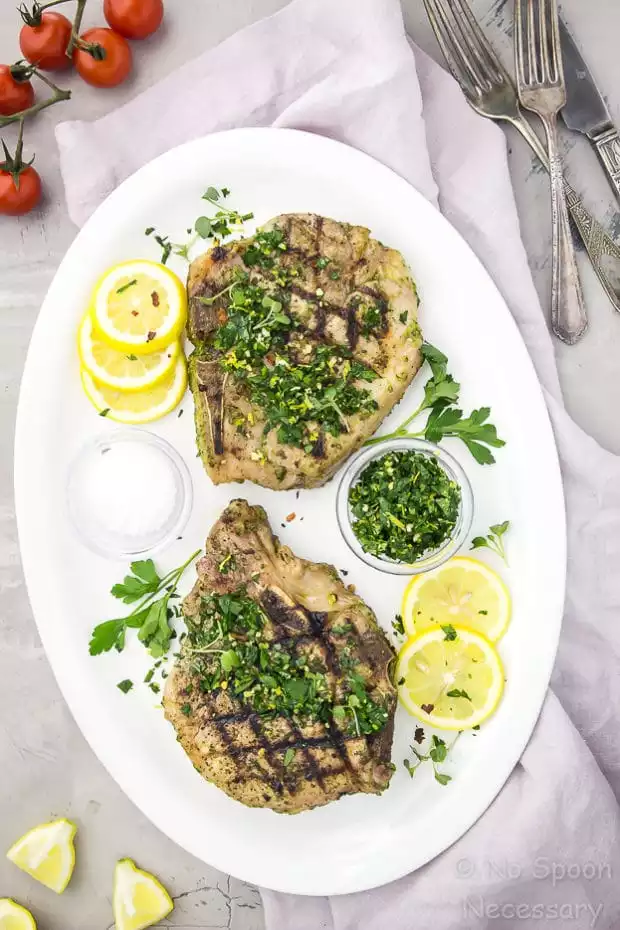 Overhead shot of two Garlic & Herb Pork Chops topped with Gremolata on a white platter with lemon slices and fresh parsley; with a pale purple napkin under the platter and vine ripe tomatoes, forks and lemon wedges surrounding the platter.