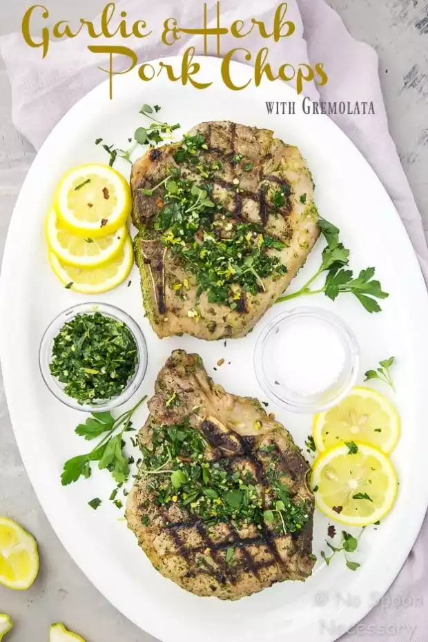 Overhead shot of two Garlic & Herb Pork Chops topped with Gremolata on a white platter with lemon slices and fresh parsley; with a pale purple napkin under the platter.