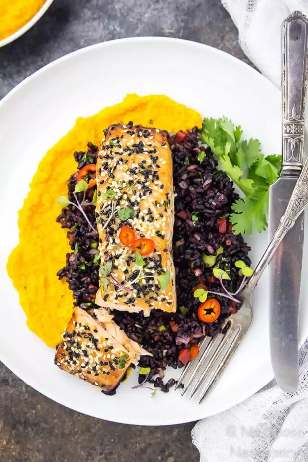 Overhead shot of cut into Honey Sesame Salmon & Asian Black Rice Salad with carrot-ginger sauce on a white plate garnished with a knife and fork.
