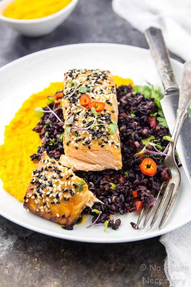 Angled shot of cut into Honey Sesame Salmon & Asian Black Rice Salad with carrot-ginger sauce on a white plate with a knife and fork.