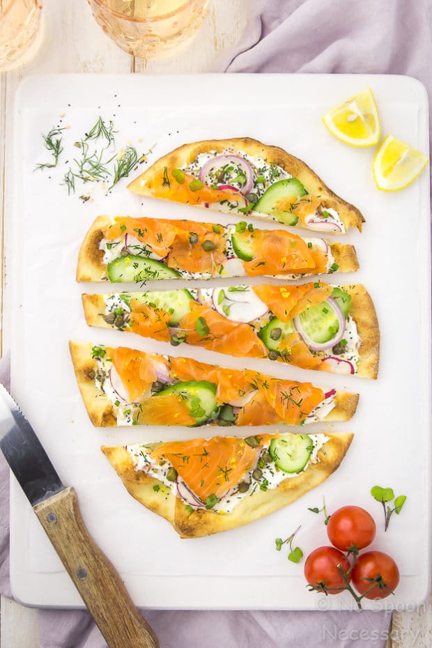 Overhead shot of Loaded Spring Salmon Pizza on a white surface with a wooden handled knife, fresh dill, lemon wedges, vine ripe tomatoes and two peach colored water glasses surrounding the pizza.