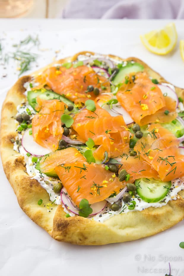 Overhead shot of Loaded Spring Salmon Pizza on a white surface with a fresh dill, lime wedges and a pale purple linen blurred in the background.