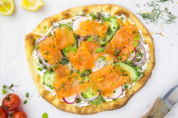 Overhead, landscape shot of Loaded Spring Salmon Pizza on a white surface with a wooden handled knife, fresh dill and vine ripe tomatoes surrounding the pizza.