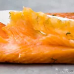 Straight on, landscape photo of a filet of salmon gravlax with a slicing knife and one single, thinly sliced piece of gravlax laying on top of the filet.