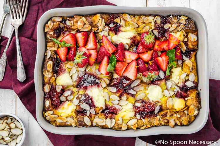 Overhead shot of Overnight Strawberry & Ricotta Breakfast Strata in a gray baking dish on a red linen with a small bowl of sliced almonds and forks surrounding the baking dish.