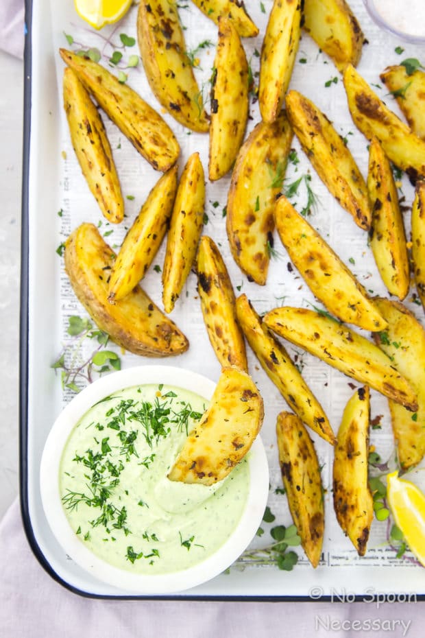 Overhead shot of a white tray of Crispy Baked Greek Potato Wedges with a potato wedge dipped in a bowl of Whipped Herbed Feta; the potatoes are garnished with fresh herbs and lemon wedges.