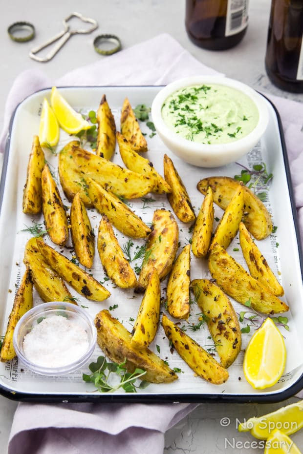 45 degree angle shot of a white tray of Crispy Baked Greek Potato Wedges with a bowl of Whipped Herbed Feta, ramekin of salt, lemon wedges and fresh herbs with beer bottles, caps and a bottle opener blurred in the background.