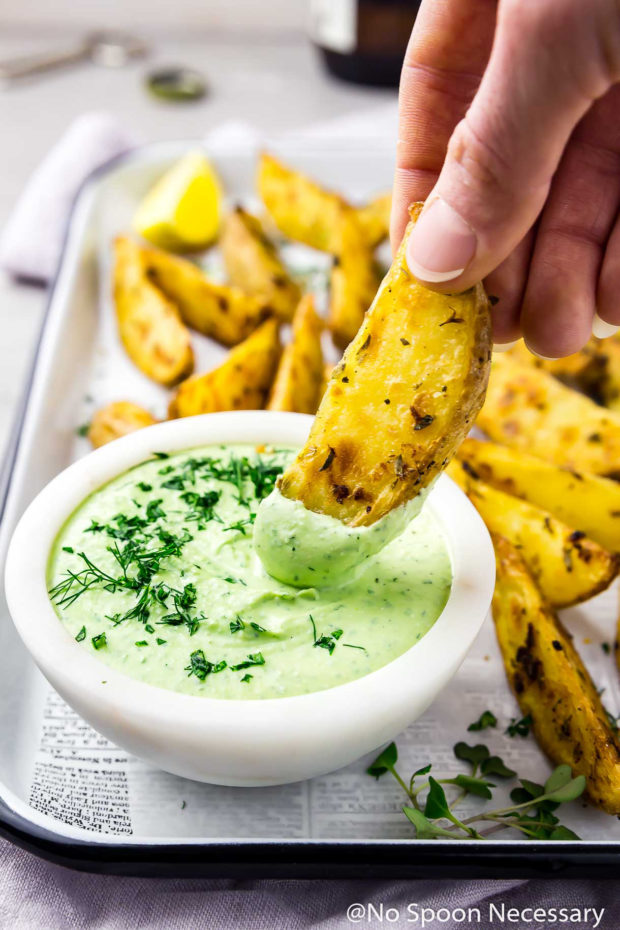 45 degree angle shot of a hand holding a Crispy Baked Greek Potato Wedge and dipping it into a bowl of Whipped Herbed Feta; with more potato wedges, beer bottle, cap and opener blurred in the background.