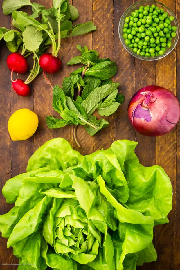 Overhead photo of a bowl of peas, head of lettuce, red onion, mint leaves, radishes, and a lemon on a kitchen counter.