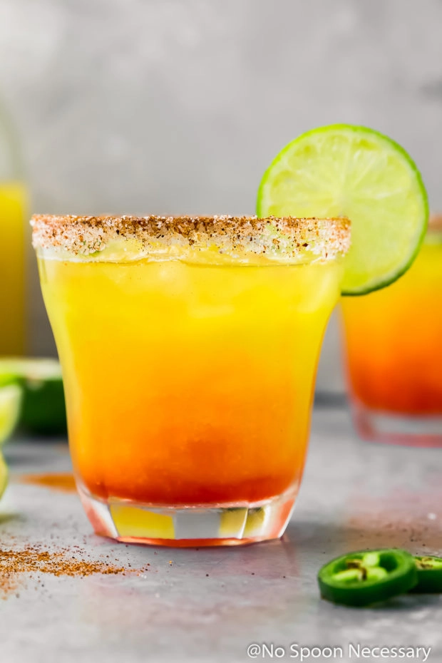 Straight on photo of a spicy mango margarita on the rocks garnished with a chili salt rim and a wheel of lime in a cocktail glass.