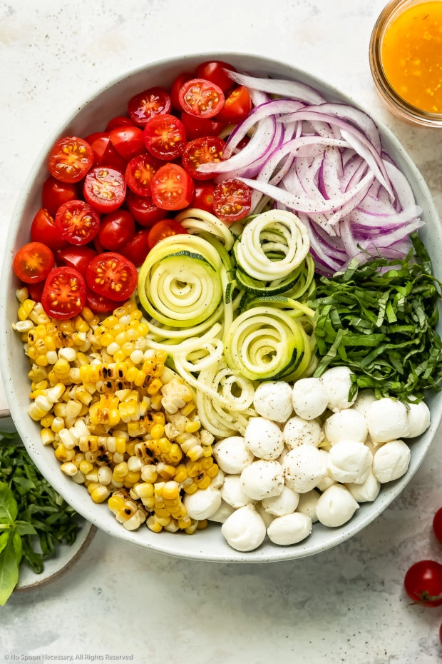 Overhead photo of all the ingredients in zucchini salad recipe neatly organized by individual ingredient in a large bowl with a jar of Italian dressing and ramekin of fresh basil next to the bowl.