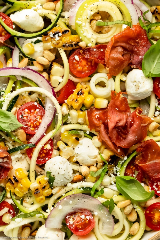 Overhead close-up photo of Italian Zucchini Salad topped with corn, tomatoes, mozzarella pearls and crispy prosciutto.