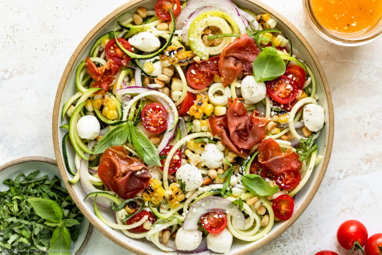 Overhead photo of Zucchini Salad topped with corn, tomatoes, mozzarella pearls and crispy prosciutto in a large white serving bowl with a jar of homemade Italian dressing and ramekin of fresh basil next to the bowl.