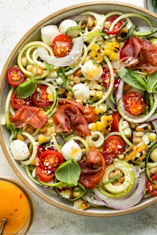 Overhead photo of zucchini spaghetti salad topped with corn, tomatoes, mozzarella pearls and crispy prosciutto and drizzled with Italian dressing in a large white serving bowl.