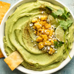 Overhead landscape photo of Spicy Avocado Hummus topped with charred corn, crumbled cheese, lime wedges and fresh cilantro in a large white serving bowl with pita strips and a ramekin of sriracha powder next to the bowl.