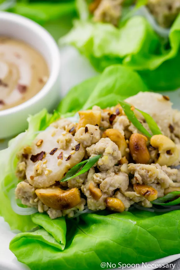 45 degree angle shot of a Cashew Chicken Lettuce Wraps on a white platter with a small ramekin of tahini sauce and more lettuce wraps partially visible and blurred in the background.
