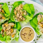 Overhead, landscape shot of Cashew Chicken Lettuce Wraps on a white platter with a small ramekin of tahini sauce on a light blue linen with a small ramekin of sliced scallions next to the platter.