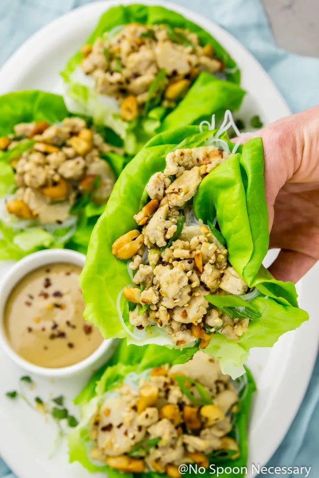 Overhead shot of a hand holding a Cashew Chicken Lettuce Wrap over a white platter of more lettuce wraps with a small ramekin of tahini sauce on a light blue linen.