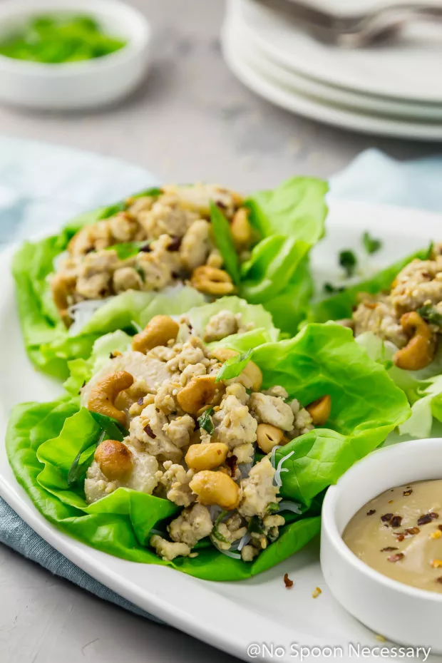 45 degree angle shot of Cashew Chicken Lettuce Wraps on a white platter with a small ramekin of tahini sauce on a light blue linen with a small ramekin of sliced scallions, a stack of plates and forks blurred in the background.
