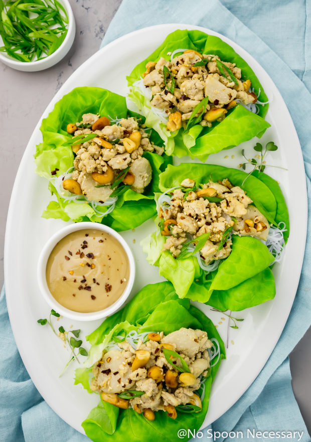 Overhead shot of Cashew Chicken Lettuce Wraps on a white platter with a small ramekin of tahini sauce on a light blue linen with a small ramekin of sliced scallions next to the platter.