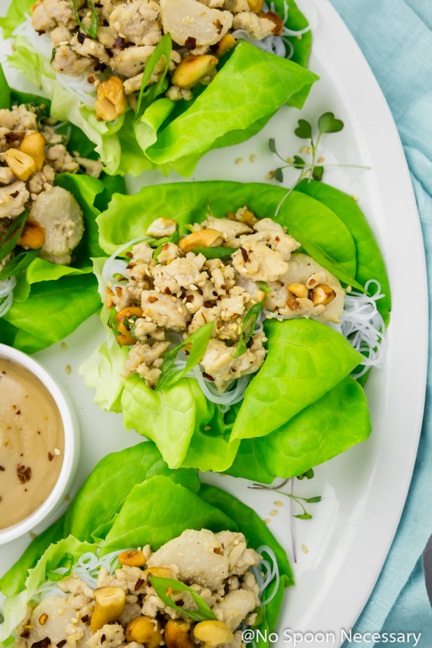 Overhead shot of Cashew Chicken Lettuce Wraps on a white platter with a small ramekin of tahini sauce on a light blue linen, with the focus on one individual wrap.