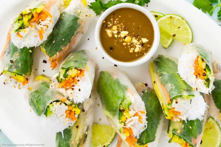 Overhead photo of Chicken Rice Paper Rolls with slices of lime and a small ramekin of peanut sauce on a large white serving platter.