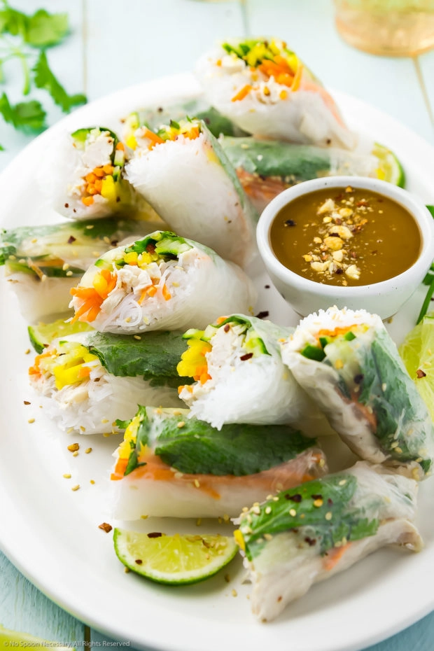 Angled photo of Chicken Rice Paper Rolls with slices of lime and a small ramekin of peanut sauce on a large white serving platter. 
