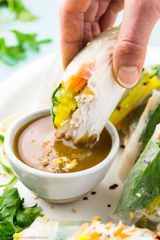 Angled photo of a Chicken Summer Roll being gripped by a person's hand and dunked into a small bowl of peanut sauce.