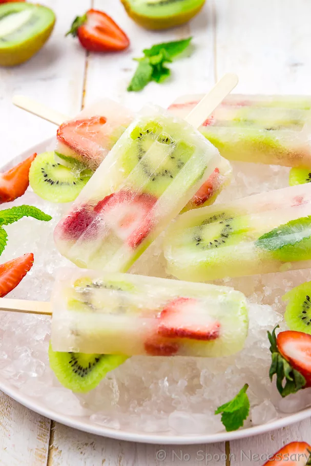 Angled shot of a pink plate of Strawberry Kiwi Mojito Boozy Popsicles on ice with strawberries, kiwis and mint in the background