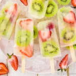 Overhead, landscape shot of Strawberry Kiwi Mojito Boozy Popsicles on a pink plate lined with ice cubes with fresh strawberries and sliced kiwis strewn about the shot.