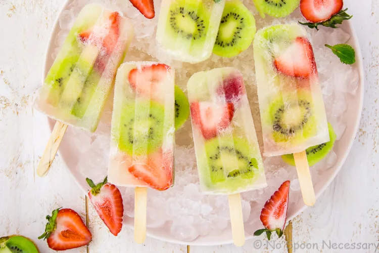 Overhead, landscape shot of Strawberry Kiwi Mojito Boozy Popsicles on a pink plate lined with ice cubes with fresh strawberries and sliced kiwis strewn about the shot.