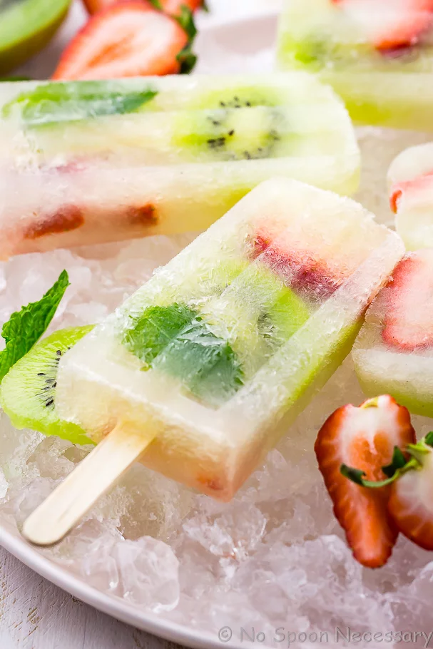 Angled close up shot of a Strawberry Kiwi Mojito Boozy Popsicle with other popsicles, strawberries, kiwi and mint blurred in the background and forefront
