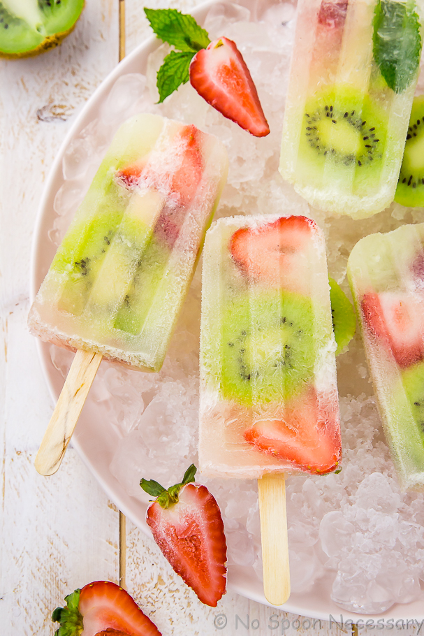 Overhead shot of half a plate of Strawberry Kiwi Mojito Boozy Popsicles on ice with sliced strawberries and mint leaves.