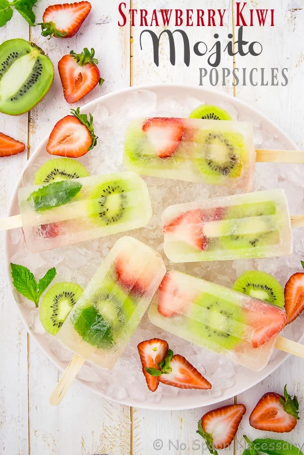 Overhead shot of Strawberry Kiwi Mojito Boozy Popsicles on a pink plate with ice, sliced strawberries, kiwis and fresh mint leaves.