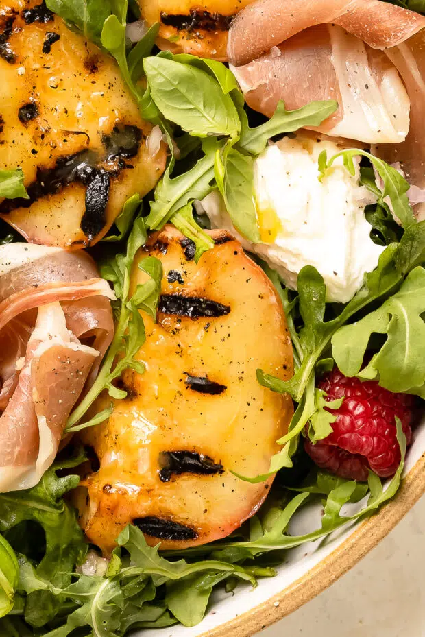Overhead, close-up photo of a torn piece of burrata and a grilled peach on a bed of salad.
