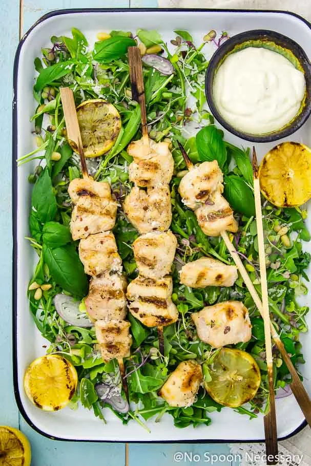Overhead of a tray of Grilled Honey Lemon Chicken Skewers on a bed of Arugula and Basil Salad with a bowl of lemon yogurt dip and lemon wedges